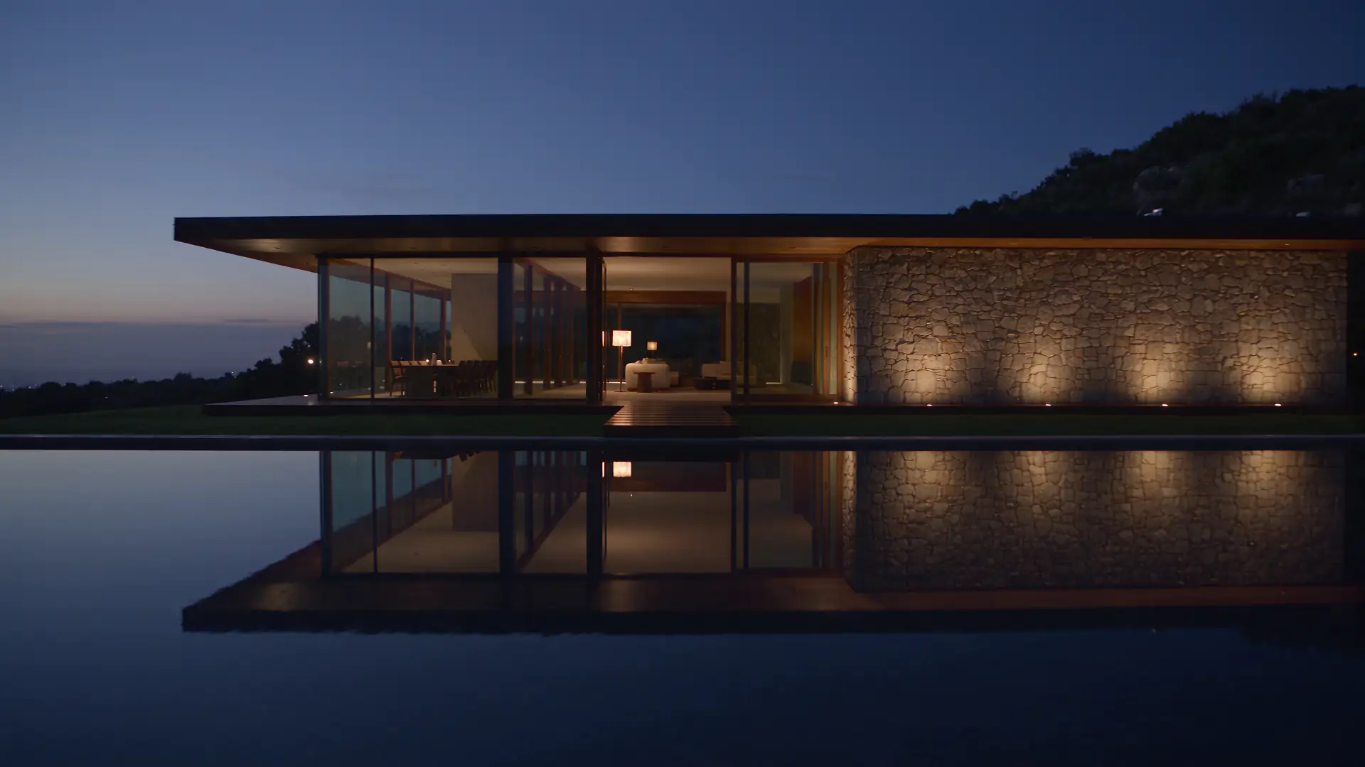 Glass house with a lit stone wall across a reflecting pool at dusk